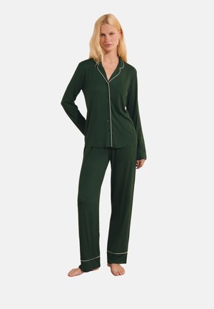 Donna bionda in piedi a piedi nudi che indossa un pigiama verde scuro a maniche lunghe con bottoni e piping bianco sui bordi e sui polsini.