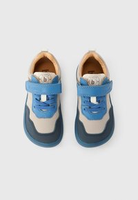 Ζευγάρι από μπεζ και μπλε παιδικά πάνινα παπούτσια με αυτοκόλλητα λουράκια και κορδόνια, φαίνονται από πάνω σε απλό φόντο.