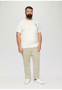 Witte katoen t-shirt met blauwe grafische print, beige corduroy broek en witte sneakers, staand op een neutrale achtergrond.