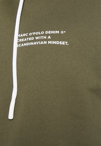Olivgrüner Sweatshirt mit weißem Kordelzug. Der Text lautet "MARC O’POLO DENIM® MIT EINEM SKANDINAVISCHEN DENKEN GESTALTET." Weicher Stoff.