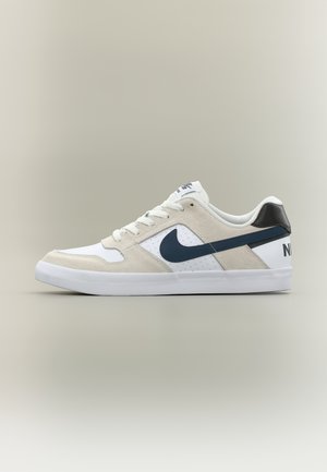 Białe i beżowe niskie sneakers Nike z granatowym logo i czarnym zapiętkiem, pokazane z profilu na neutralnym tle.