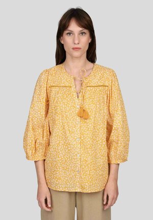 Blouse jaune avec un motif floral ; présente des manches bouffantes, un devant boutonné et un détail à nouer au niveau du col avec une accent de pompon.