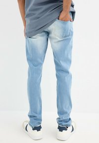 Jeans de denim azul claro con un corte ajustado, que presentan un patrón desgastado y dos bolsillos traseros. Combinados con zapatillas blancas y azul marino.