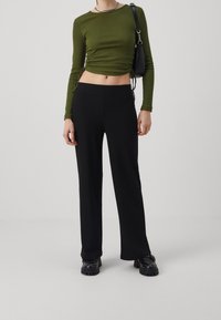 Vrouw in een olijfgroene lange mouw ruched crop top, zwarte geribbelde wijde broek, zwarte gespte schoenen en met een zwarte schoudertas.