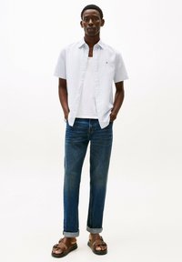 Camisa de botón de manga corta, a rayas de color azul claro, camiseta blanca debajo, vaqueros oscuros, sandalias marrones con detalles de tiras, corte relajado.