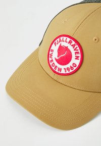 Căciulă bej din material moale, cu o margine curbată. Prezintă o emblemă circulară roșie și albă cu un logo de vulpe și textul "FJÄLLRÄVEN SWEDEN 1960".