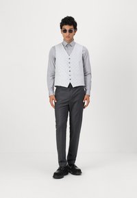 Helles graues gemustertes Weste über einem knopfgeknöpften grauen Hemd, kombiniert mit dunkelgrauen maßgeschneiderten Hosen und schwarzen Schuhen. Runde Sonnenbrillen runden den Look ab.