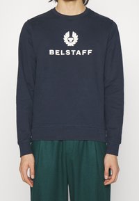 Marinblå Belstaff-tröja med vitt logotyp och text på bröstet, bärs av en person med gröna byxor synliga från midjan och ner.