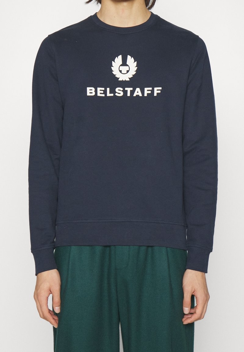 Marinblå Belstaff-tröja med vitt logotyp och text på bröstet, bärs av en person med gröna byxor synliga från midjan och ner.