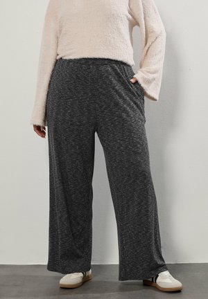 Person iført mørkegrå vidde benbukser, cremefarvet, blød langærmet top, hvide sneakers med sorte striber, stående op ad en hvid væg.