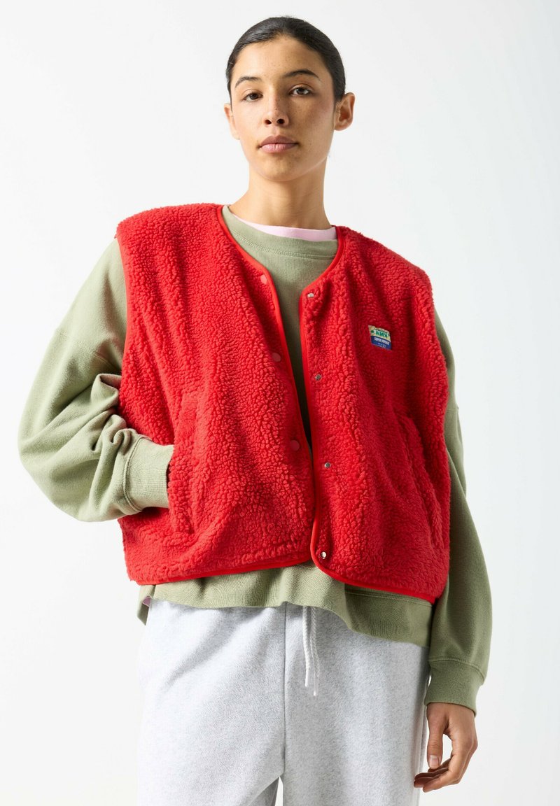 Rotes Fleece-Weste mit strukturierter Oberfläche, Druckknöpfen und Seitentaschen, über einem grünen Sweatshirt getragen, vor einem schlichten Hintergrund.