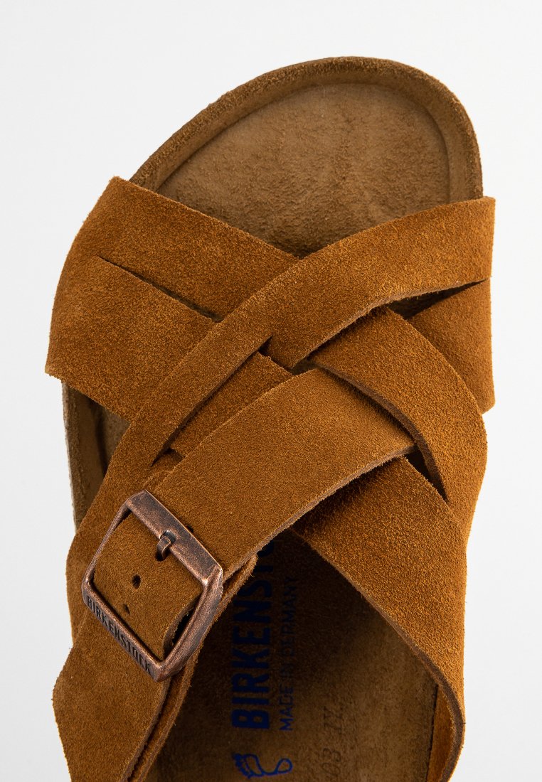Sandale en daim marron avec des brides entrecroisées, dotée d'une boucle en métal. La semelle intérieure est douce et texturée, marquée du nom de la marque en bleu.