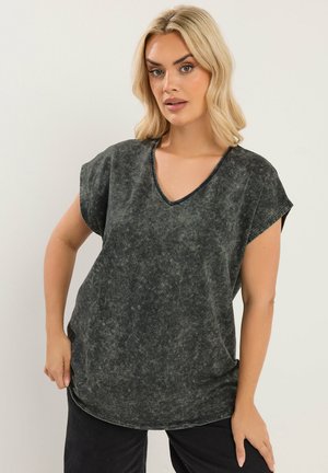 Lyshåret kvinde iført en løs, sort syrevaske V-hals T-shirt og sorte bukser, stående mod en ensartet lys baggrund.