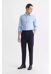 Jasnoniebieska koszula z długim rękawem z guzikami połączona z ciemnymi, dopasowanymi spodniami; wyróżnia się prostym krojem i eleganckimi czarnymi butami.