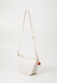 Weiße Handtasche aus Kunstleder mit trapezförmiger Form, verstellbarem Tragegurt, goldfarbenen Beschlägen und einem dekorativen Quast mit einer braunen Perle.