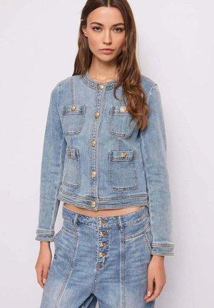 Joven mujer con chaqueta vaquera azul clara con botones dorados y jeans de tiro alto a juego con cierre de botones, posando contra un fondo sencillo.