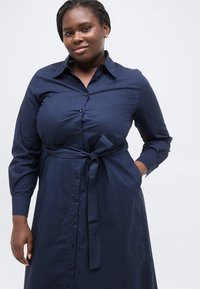 Γυναίκα με πλεξούδες, φορούσα μπλε φορεμα με κουμπιά και ζώνη δεμένη στη μέση, στέκεται με το ένα χέρι στην τσέπη της μπροστά σε ένα λευκό φόντο.