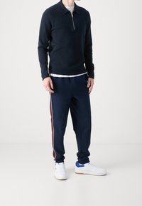Marinblå zip-up stickad tröja med krage, kombinerad med marinblå sweatpants med vinröda sidostripes och vita sneakers med blå detaljer.