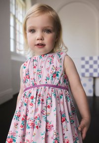 Rožinė medvilninė gėlių rašto suknelė su purpuriniu liemens diržu; turi trumpas rankoves, pūstą dizainą ir daugių spalvų gėlių raštus.