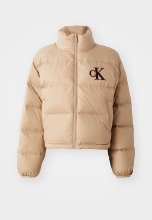 Zkrátená puffová bunda v béžovej farbe, s vysokým golierom, zipsom a hnedým vyšívaným logom "ck" na hrudi. Vyrobená z jemného, polstrovaného materiálu.
