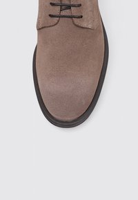 Zapato de ante marrón con punta redonda y cordones negros, que presenta una suela negra en contraste y una textura suave. Vista desde arriba.