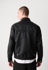 Veste à carreaux noirs avec col montant et logo brodé au dos, dotée d'une texture lisse et de détails à boutons-pression.