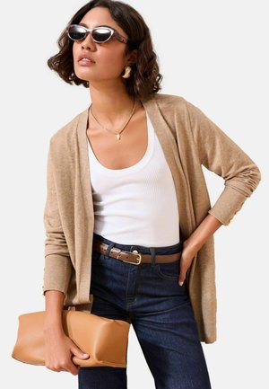 Cardigan bej cu mâneci lungi, purtat peste un top din bumbac alb cu dungi. Blugi albaștri, curea maro și o poșetuță bej. Ochelari rotunzi completează ținuta.