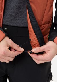 Geïsoleerde oranje jas met zwarte accenten heeft een verstelbare trekkoordtaille; handen trekken de zwarte koord eronder strakker. Draagt over aansluitende zwarte broek.