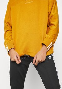 Gul sweatshirt med strukturerad yta, som har rund halsringning och vita tre-stripes detaljer vid ärmarna. Svarta byxor med vita sidostripes.