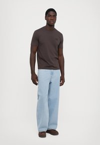 Tricou din bumbac de culoare maro închis, cu mâneci scurte, cu un logo negru, asortat cu blugi largi de culoare albastru deschis și teniși maronii.
