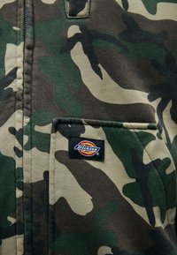 Kameržaudens jaka ar zaļiem, brūniem un beidzīgiem rakstiem. Iekļauj priekšējo kabatu ar melnu "Dickies" logo uzšuvumu.