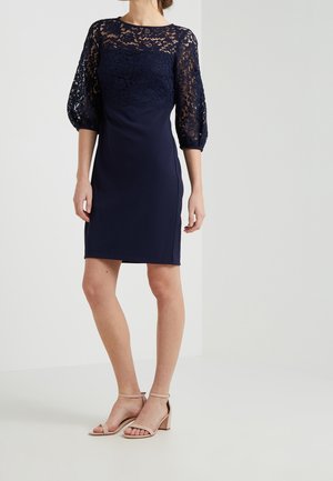 Robe de marine avec un haut en dentelle et des manches bouffantes, jupe lisse et ajustée, accompagnée de sandales à talons blocs nude, sur fond clair.