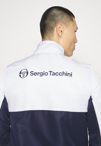Balta un tumši zilā jaka ar augstu apkakli, kuras aizmugurē ir liels "Sergio Tacchini" logo. Izgatavota no gluda, viegla materiāla.
