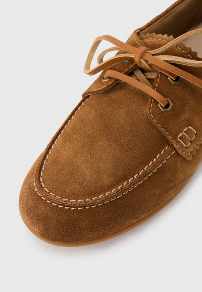 Καφέ μοκασίνι από σουέντ με ορατή λευκή ραφή και δερμάτινες κορδόνια δεμένα σε φιόγκο σε ανοιχτό φόντο.