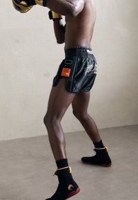 Schwarze Boxershorts mit einem verstellbaren Bund, die orangefarbene und weiße Akzente sowie seitliche Schlitze aufweisen. Schwarze Schuhe mit gelben Details.