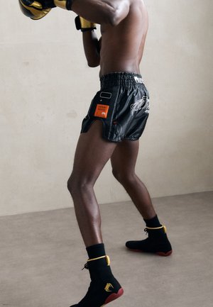 Black boxing shorts with a drawstring waistband, featuring orange and white accents and side slits. Black shoes with yellow details.