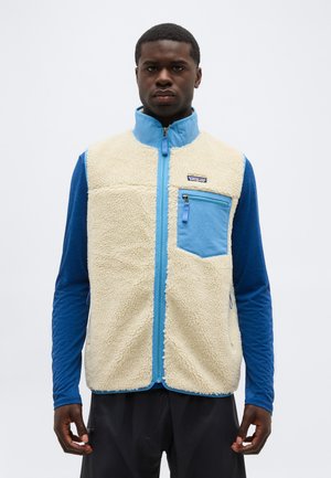 Vyras dėvi kreminį vilnos liemenę su mėlynu užtrauktuku ir krūtinės kišene ant ilgomis rankovėmis mėlynos spalvos marškinių, stovintis priešais vientisą foną.