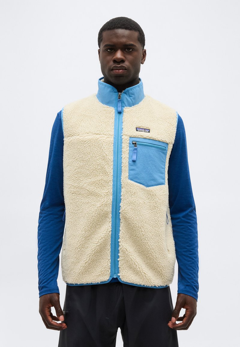 Vīrietis valkā krēmkrāsas vilnas vestu ar zilu rāvējslēdzēju un krūšu kabatu virs zilas garroku krekla, stāvot pret vienveidīgu fonu.