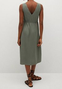 Oliwkowa sukienka midi z plecami w kształcie litery V, elastycznym pasem i zwiewną spódnicą. Połączona z czarnymi płaskimi sandałami sznurowanymi.