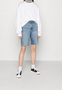 Pantalones cortos de mezclilla azul claro con dobladillo deshilachado, combinados con una blusa blanca de manga larga, zapatillas blancas y negras, y calcetines blancos. Diseño sencillo.