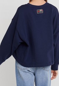 Marinblå oversized sweatshirt med ribbad halsringning och kontrasterande lappdetalj på övre ryggen. Mjukt, texturerat tyg, avslappnad passform.