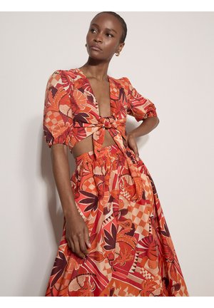 Kobieta w pomarańczowo-czerwonym kwiecistym dwuczęściowym stroju z bufkami na rękawach, topem z węzłem z przodu i spódnicą z wysokim stanem na jednolitym tle.