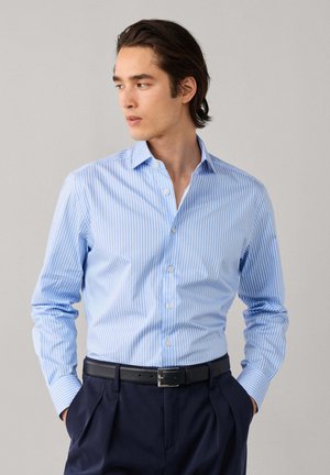 Homme portant une chemise à manches longues rayée bleu clair et blanc, un pantalon bleu marine foncé et une ceinture noire, regardant vers sa gauche avec les mains dans les poches.