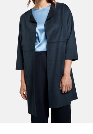 Vrouw draagt een loszittende, open zwarte jas met driekwart mouwen over een lichtblauw satijnen topje en zwarte broek.