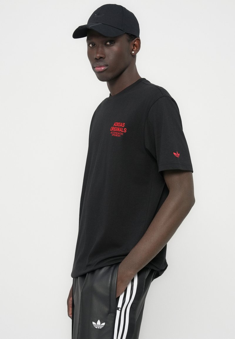 T-shirt en coton noir avec logo "Adidas Originals" rouge sur la poitrine. Porté avec un pantalon noir présentant des bandes latérales blanches et le logo Adidas.