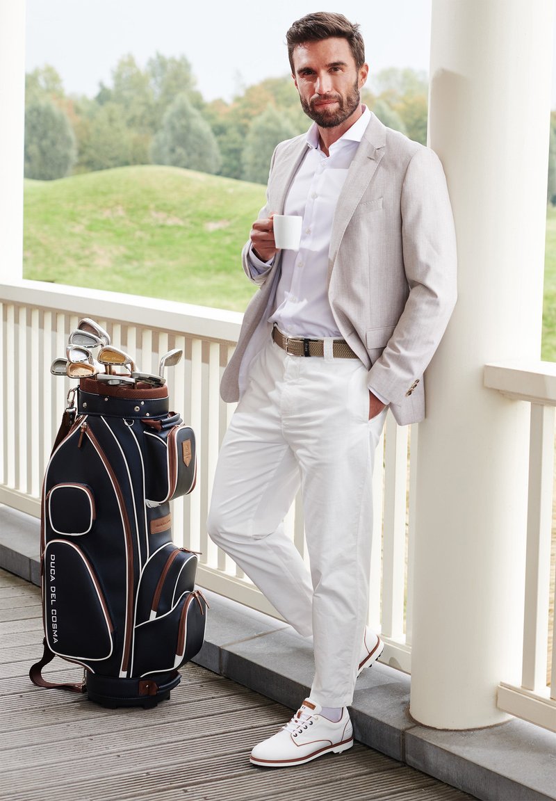 Golf bag in navy with brown accents, featuring multiple pockets and clubs. Model wears a light blazer, white shirt, and white shoes.