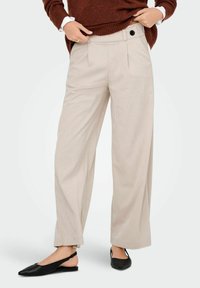 Pantalon large rayé beige avec une taille haute, des poches latérales et un détail en bouton noir, associé à des ballerines noires à l'arrière ouvert.