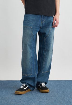 Person trägt blaue Jeans mit weitem Bein und großen aufgesetzten Taschen vorne sowie schwarze Turnschuhe mit weißen Schnürsenkeln und Gummisohlen auf blauem Teppich.