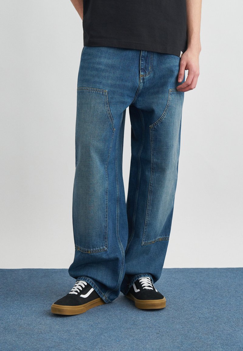 Persona con vaqueros azules de pierna ancha y grandes bolsillos delanteros de parche, y zapatillas negras con cordones blancos y suelas de goma, sobre una alfombra azul.