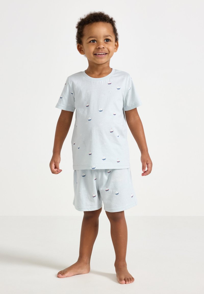Completo pigiama in cotone azzurro chiaro, composto da una maglietta a maniche corte e pantaloncini, decorato con piccoli motivi di barche in rosso e blu.